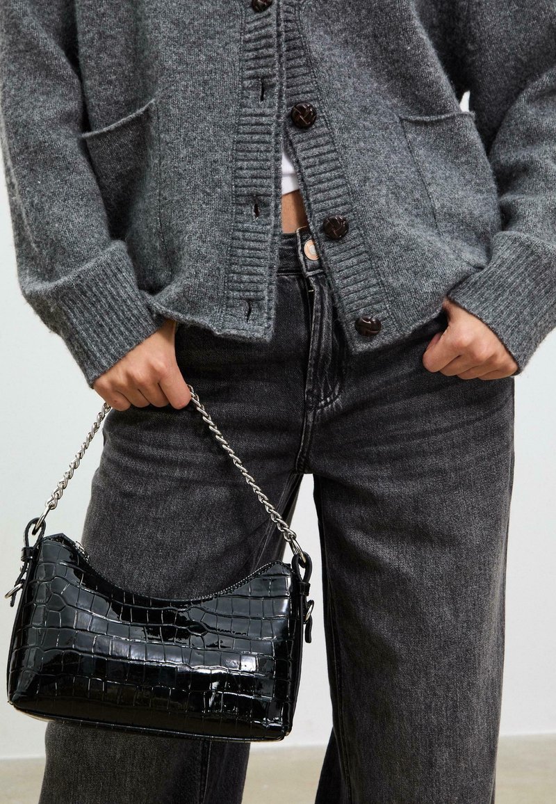 Sort patenteret læderhåndtaske med krokodillemønster, udstyret med en sølv kædrestropp. Bæres med en grå cardigan og mørke jeans.