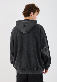 Hoodie negru de mari dimensiuni, realizat dintr-un material moale și texturat. Include o glugă și detalii decorative cu ținte argintii pe mâneca stângă. Finisaj uzat.