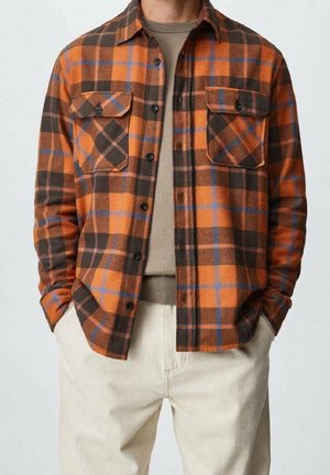 Hombre vestido con camisa de cuadros naranja, marrón y azul con botones, encima de un jersey beige y pantalones claros, manos en los bolsillos.