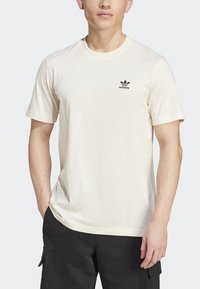 Béžové tričko s krátkým rukávem vyrobené z bavlny, s černým logem Adidas na levé části hrudi, volný střih a kulatý výstřih.