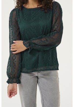 Mujer con una blusa de encaje verde oscuro de manga larga con patrones de diamante y jeans gris claro, con las manos cruzadas suavemente a la altura de la cintura.