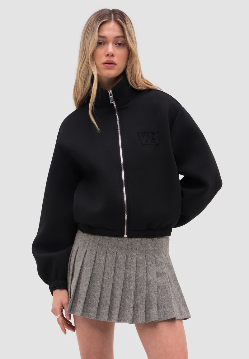 Nuori nainen, jolla on pitkät vaaleat hiukset, pukeutuneena mustaan vetoketjulliseen takkiin, jossa on WB-logo, ja harmaaseen laskostettuun minihameeseen.