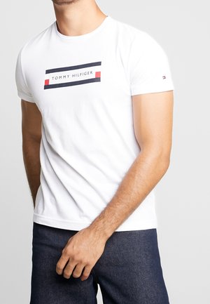 Mężczyzna w białej koszulce Tommy Hilfiger z granatowym i czerwonym logo na klatce piersiowej oraz ciemnoniebieskich dżinsach na jednolitym tle.