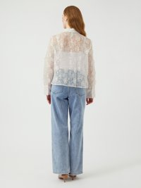 Blusa in pizzo floreale bianco trasparente con maniche lunghe, colletto e vestibilità ampia, abbinata a jeans a gamba larga blu chiaro.