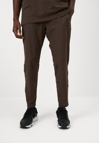 Pantalons de sport athlétiques bruns, ajustés et en matériau léger, dotés de poches latérales et d'un logo discret. Portés avec des chaussures de sport noires.