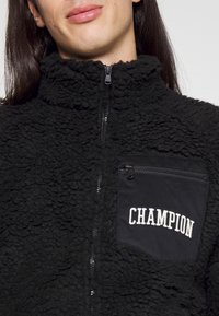 Sort fleecejakke med en tekstureret, fluffy overflade, høj krave og en lynlåslomme med "CHAMPION" broderi i hvid.