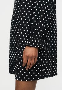 Χέρι που ακουμπά δίπλα σε μαύρο φόρεμα με λευκές πουά και ελαστική μανσέτα με βολάν στο μανίκι.