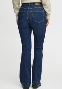 Vaqueros acampanados de color azul oscuro, fabricados en denim, con dos bolsillos traseros y una etiqueta en la cintura. La tela tiene una apariencia lisa, con una textura ligeramente rugosa.