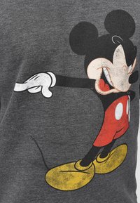 Mickey Mouse portant un short rouge et des chaussures jaunes, souriant et pointant vers la gauche, imprimé sur une surface en tissu gris.
