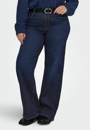 Persoon draagt donkerblauwe wijde jeans met een zwarte riem en zwarte schoenen, staat met één hand de riem verstelend.