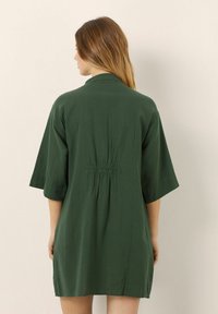 Zelené krátké šaty s volným střihem, které mají rukávy ve stylu kimono a detaily na stažení v pase, vyrobené z lehkého materiálu.