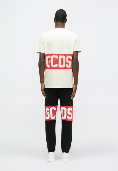 T-shirt crema a maniche corte con banda rossa "GCDS"; pantaloni neri con accenti rossi "GCDS" simili; modello che indossa sneakers bianche.