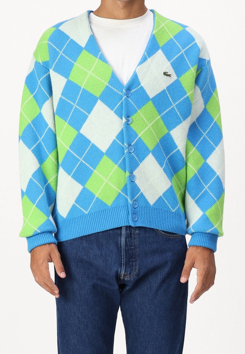 Hombre llevando un cárdigan abotonado de rombos en azul, verde y blanco sobre una camiseta blanca con pantalones vaqueros de color azul oscuro.