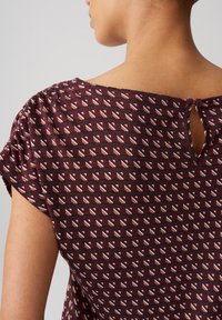 Luksus bordeaux bluse med et geometrisk mønster i creme og sort, korte ærmer og en knap-øjnehul detalje i nakken bagpå.