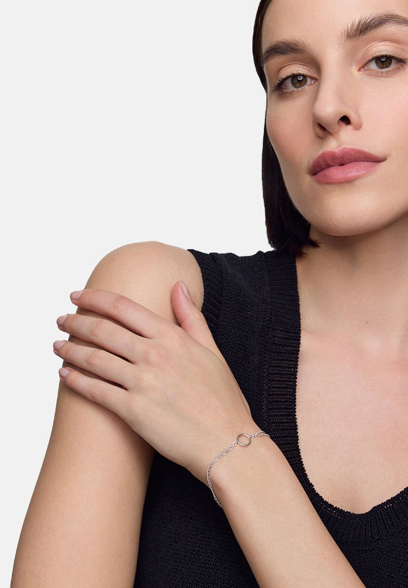 Srebrna bransoletka z okrągłym akcentem, prezentowana na nagim nadgarstku. Osoba nosząca bransoletkę ma jasną skórę i ciemne włosy, ubrana w czarną dzianinową bluzkę.