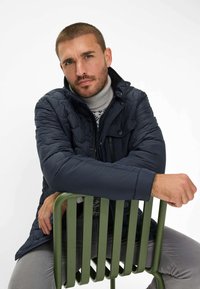 Giacca trapuntata blu navy con colletto alto, cerniera e bottoni a pressione, indossata sopra un dolcevita grigio chiaro. Sedia metallica verde sullo sfondo.