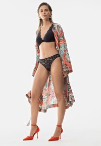 Bralette in pizzo nero e culotte coordinate abbinate a un kimono arancione a fantasia. La modella indossa scarpe con tacco rosso. Texture lisce e colori audaci.