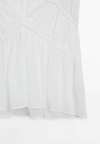 Haut blanc sans manches avec tissu froncé, présentant des coutures croisées au niveau du buste et un ourlet léger et superposé. Texture légère.