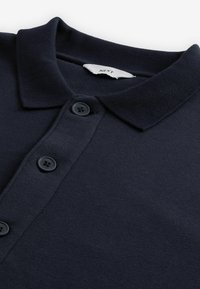 Tricou polo bleumarin cu o textură moale, guler cu dungi și placket cu trei nasturi negri; are un etichetă țesută la guler.