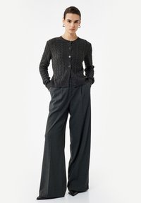 Mujer con un cárdigan de punto trenzado gris oscuro abotonado, pantalones de pierna ancha en color carbón y zapatos negros de punta, de pie con las manos en los bolsillos.