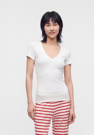 Jeune femme aux cheveux noirs portant un t-shirt blanc à col en V et un pantalon rayé rouge et blanc, debout devant un fond clair uni.