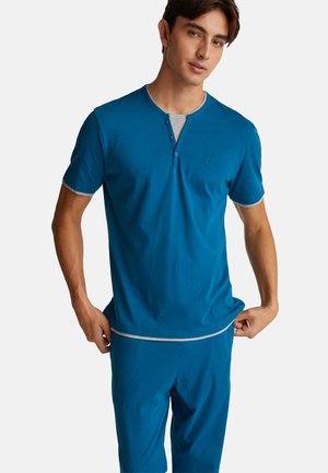 Joven vestido con una camiseta henley de manga corta azul sobre una camiseta interior gris y pantalones azules a juego, de pie contra un fondo blanco.