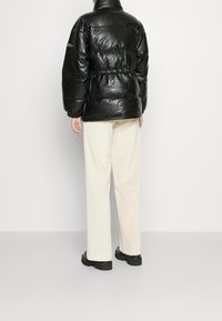 Czarna puchowa kurtka o błyszczącej fakturze, z wysokim kołnierzem i taliowaniem, zestawiona z kremowymi sztruksowymi spodniami i czarnymi butami.