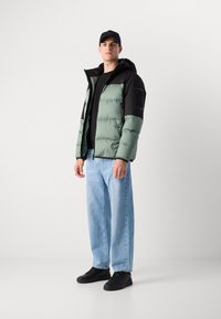 Opblaasjas in lichtgroen met zwarte accenten, voorzien van een capuchon en ritssluiting. Gecombineerd met lichtblauwe denim en zwarte sneakers.