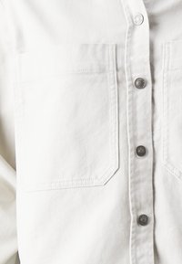 Camisa de mezclilla blanca con cierre de botones en la parte delantera, dos bolsillos en el pecho con solapas inclinadas y acentos de botones metálicos. Textura suave.