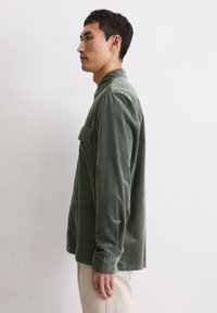 Camicia in velluto a coste verde con maniche lunghe, colletto con bottoni e una tasca sul petto. Tessuto testurizzato e vestibilità comoda.