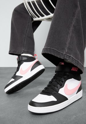Personne portant des baskets montantes Nike noires, blanches et roses avec un pantalon noir, assise sur une chaise en fil blanc sur un sol gris.