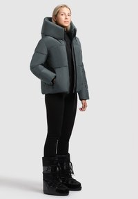 Ung kvinde iført en grå puffjakke med høj krave, sorte leggings og sorte snørede vinterstøvler, der står foran en ensfarvet baggrund.