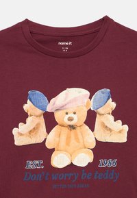 Három mackó, beretthatszban, egy bordó pólón, amelyen a felirat: "EST. 1986 Ne aggódj, légy mackó! Jobb napok előttünk!"