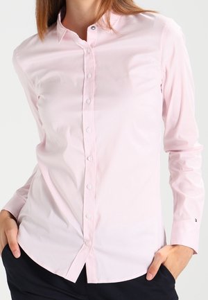 Woman wearing a light pink button-up long-sleeve shirt with a collar, hands in black pants pockets, standing against white background.