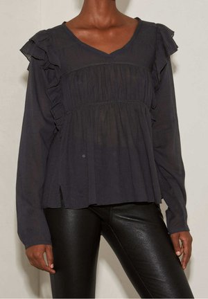 Frau trägt eine schwarze Bluse mit langen Ärmeln und Rüschendetails sowie schwarze Lederhosen, steht vor einem schlichten hellen Hintergrund.