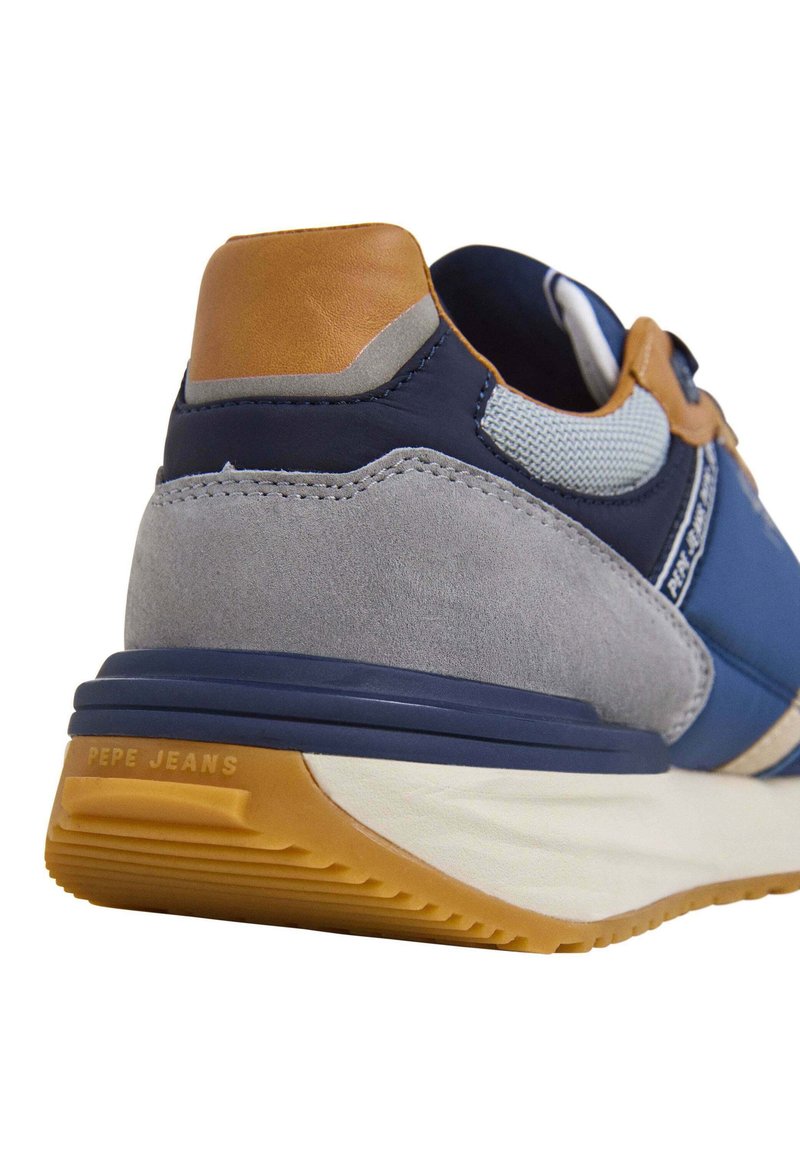 Baskets navy et gris avec un talon en daim texturé, des accents en cuir beige, des panneaux en maille, et une semelle en caoutchouc blanc et caramel. Nom de la marque visible.