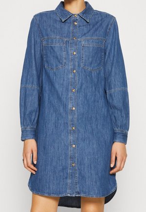 Blauwe denim overhemdjurk met lange mouwen, knoopsluiting aan de voorkant, twee borstzakken en een kraag, gedragen door een persoon.