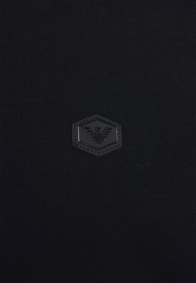 Svart tyg med en slät textur. Har en svart hexagonal logotypapplikation med en präglad design. Enkelt och minimalistiskt stil.