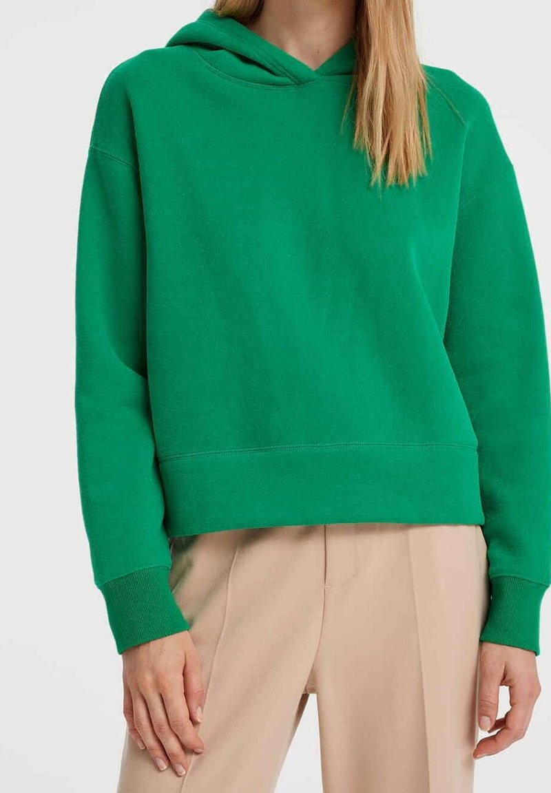 Femme portant un sweat à capuche vert vif et un pantalon beige, debout devant un fond clair uni.