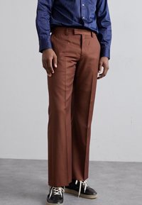 Pantalon marron avec une coupe droite, fabriqué dans un matériau lisse, assorti à des sneakers noirs. La chemise est bleue avec des rayures blanches.