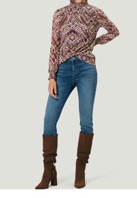 Haut à manches longues à motifs dans des teintes de rouge et de crème, associé à un jean bleu et des bottes à talons hautes en cuir marron, décontractées. Tissu texturé.