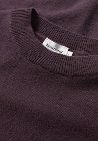 Lila Strickpullover mit rundem Ausschnitt und geripptem Saum, aus weichem Material gefertigt, mit einem Etikett, das Marke und Größe angibt.