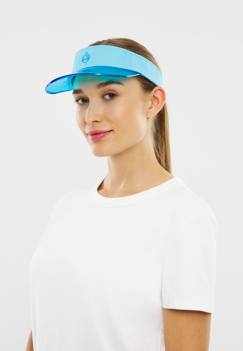 Mladá žena so svetlou pleťou a dlhými vlasmi má na hlave modrý slnečný štít a obyčajné biele tričko, s mierne nakloneným pohľadom a jemným úsmevom.