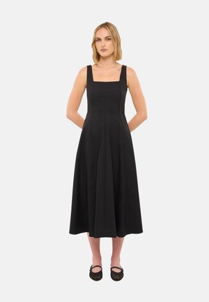 Femme portant une robe midi noire sans manches avec un décolleté carré et des chaussures plates noires, debout avec les mains derrière le dos sur un fond blanc.