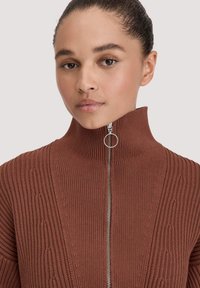 Junge Person mit Haaren im Dutt, die einen rostfarbenen gerippten Strickpullover mit hohem Kragen und silbernem Reißverschluss mit rundem Ziehstück trägt.