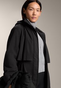 Veste extérieure noire avec capuche, présentant une coupe décontractée et une doublure intérieure texturée grise, tenue par le modèle. L'arrière-plan est neutre.