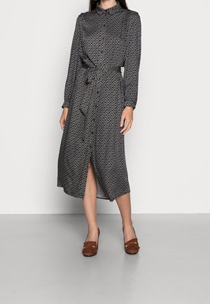 Kobieta ubrana w midi sukienkę z długim rękawem i zapinana na guziki, z geometrycznym wzorem i paskiem w talii, do której dobrała brązowe wsuwane buty na obcasie.