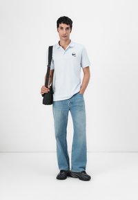 Lys blå polo skjorte med logo på brystet, kombinert med lyse jeans og svarte joggesko. Modellen holder en svart skulderveske.