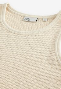 Creme strikket sweater med et tekstureret mønster, en rund halsudskæring med ribkant og et mærke med teksten "NEXT EDIT." i halsudskæringen.