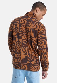 Bruine fleece jas met een patroon van abstracte ontwerpen in donkere tinten, met een kraag en lange mouwen. Lichtblauwe jeans zichtbaar.